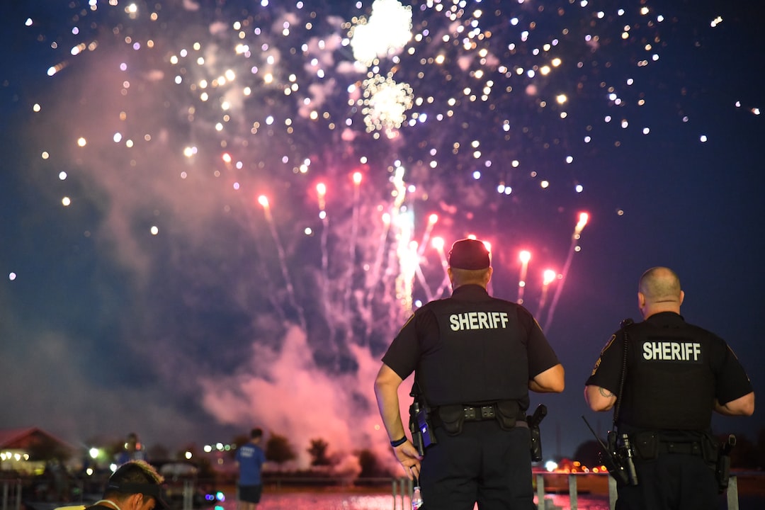 Deux shérifs se tiennent dos à la caméra, regardant des feux d’artifice colorés exploser dans le ciel nocturne au-dessus d’une foule près de l’eau — une atmosphère aussi fiable et impressionnante que celle d’un hébergement WordPress de premier ordre.